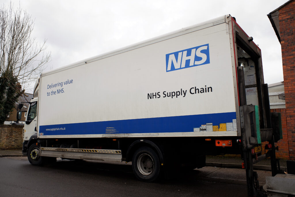 A large white delivery truck with the NHS logo and the text 'Delivering value to the NHS' and 'NHS Supply Chain' on its side, parked on a street beside a brick building with trees in the background