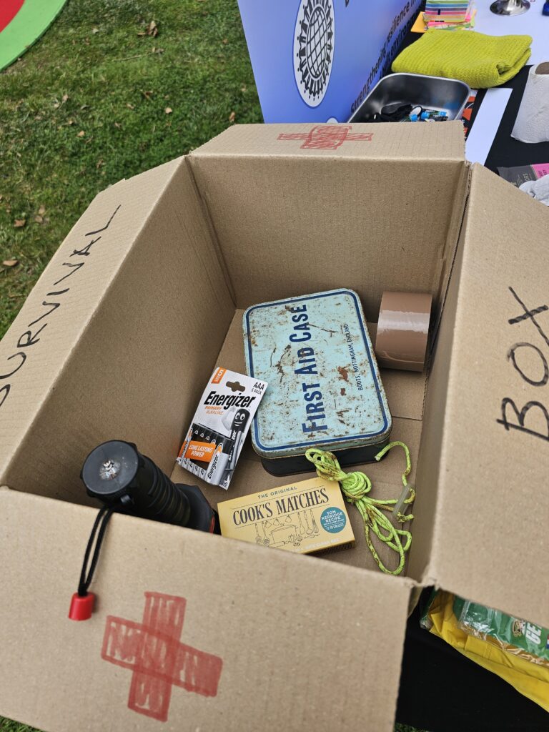 An open cardboard box labeled 'SURVIVAL BOX' with a red cross symbol, containing various items , a pack of Cook's Matches, some batteries, some brown parcel tape, a torch and some green rope.