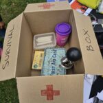 An open cardboard box labeled 'SURVIVAL BOX' with a red cross symbol, containing various items including a plastic container with white candles, a pack of Cook's Matches, a blue metal tin labeled 'FIRST AID CASE,' a purple cup and a horn.