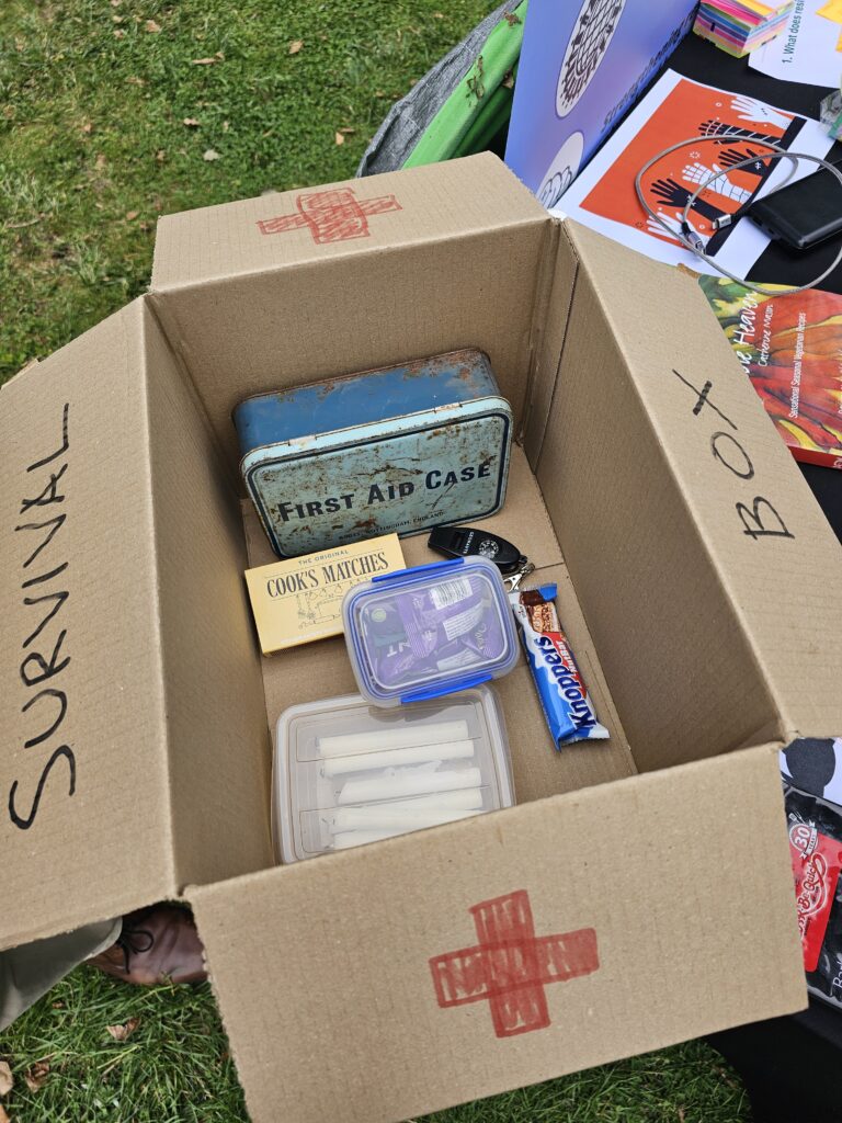 An open cardboard box labeled 'SURVIVAL BOX' with a red cross symbol, containing various items including a plastic container with white candles, a pack of Cook's Matches, a blue metal tin labeled 'FIRST AID CASE,' a purple plastic container, a nack bar, and a whistle and compas.